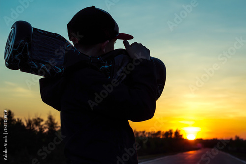 A young boy holds on his shoulder a super popular electric scooter on his shoulder against a sunset background. Mini segway hoverboard transportation device,popular city transport. 