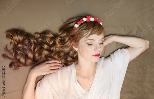 Cute dreaming girl in roses wreath and dress with curly hair lies on bed