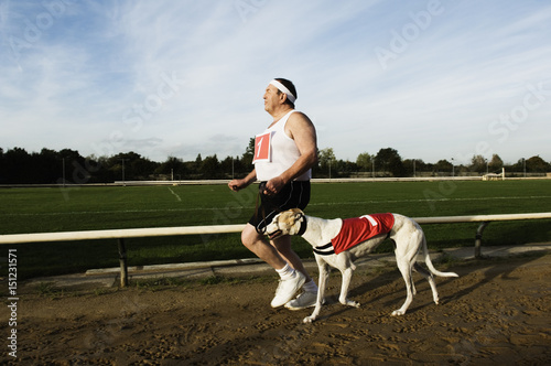 Man in sportswear running on a racetrack, with a white greyhound wearing red bib with number one.