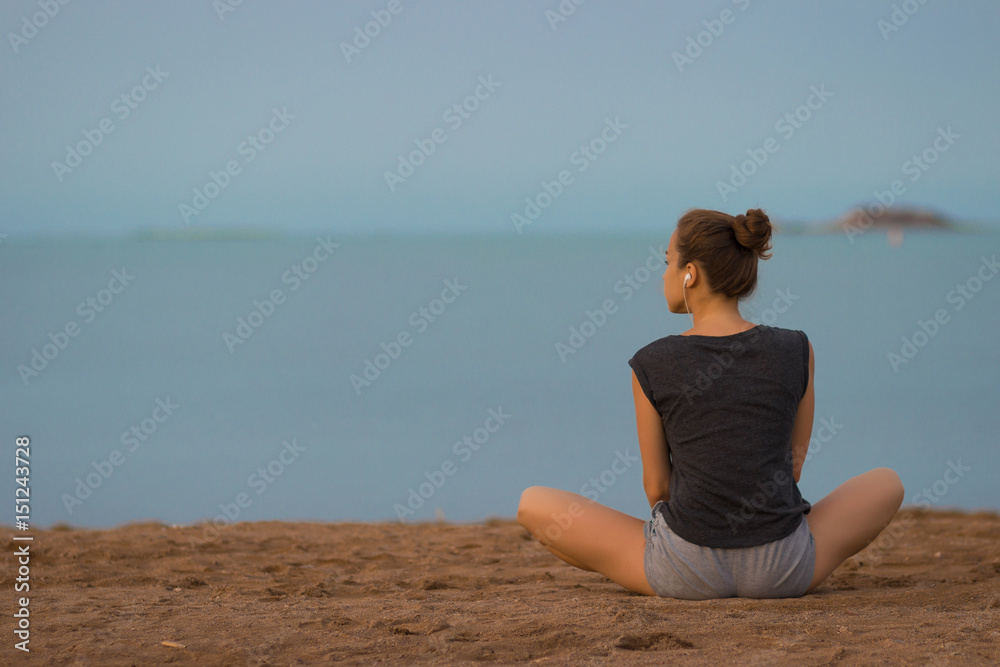 Young beautiful woman meditating on the beach in the morning at the dawn of the sun in a gray T-shirt and shorts