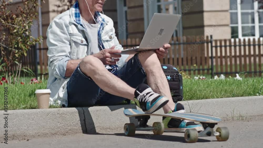 MS Lockdown of senior man with longboard sitting on sidewalk and having video call on laptop MS Lockdown of senior man with longboard sitting on sidewalk and having video call on laptop