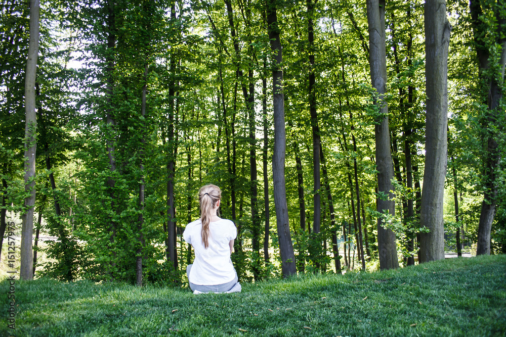 A girl in a white t-shirt sits and stares into the forest. Rear view.
