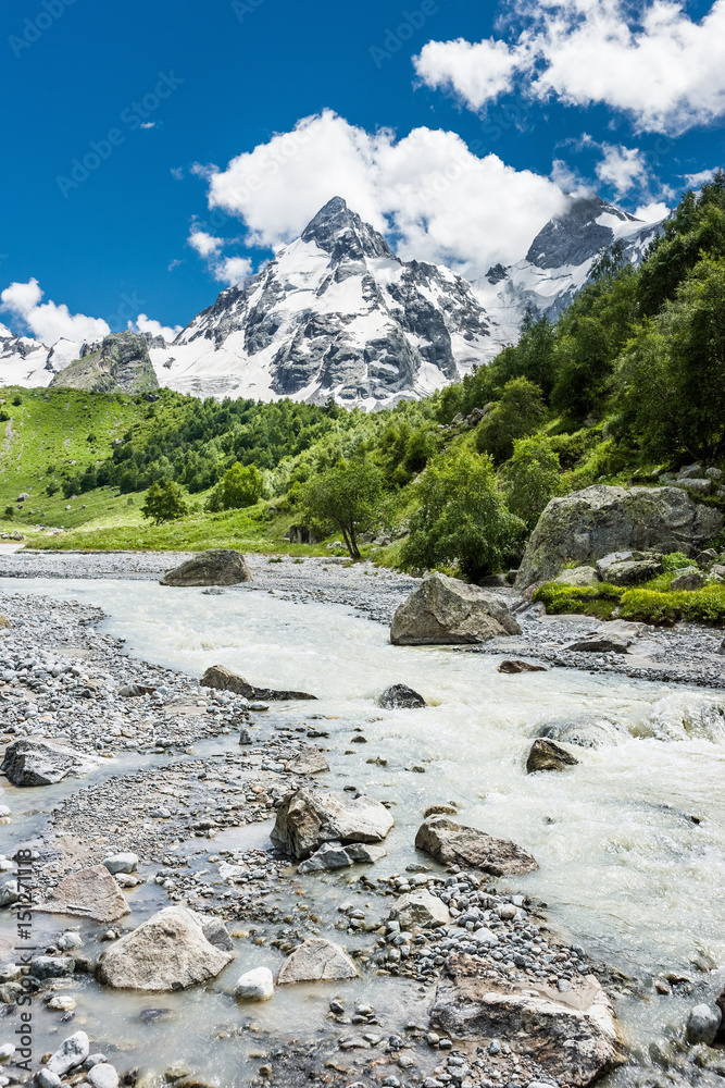Summer mountain landscape with small rough river against snowy mountains and blue sky. Russian Caucasus, Elbrus region.