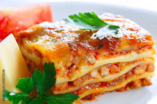 Meat lasagna. A piece of lasagna on a white plate. Next to the lasagna lies a piece of cheese and a slice of tomato.