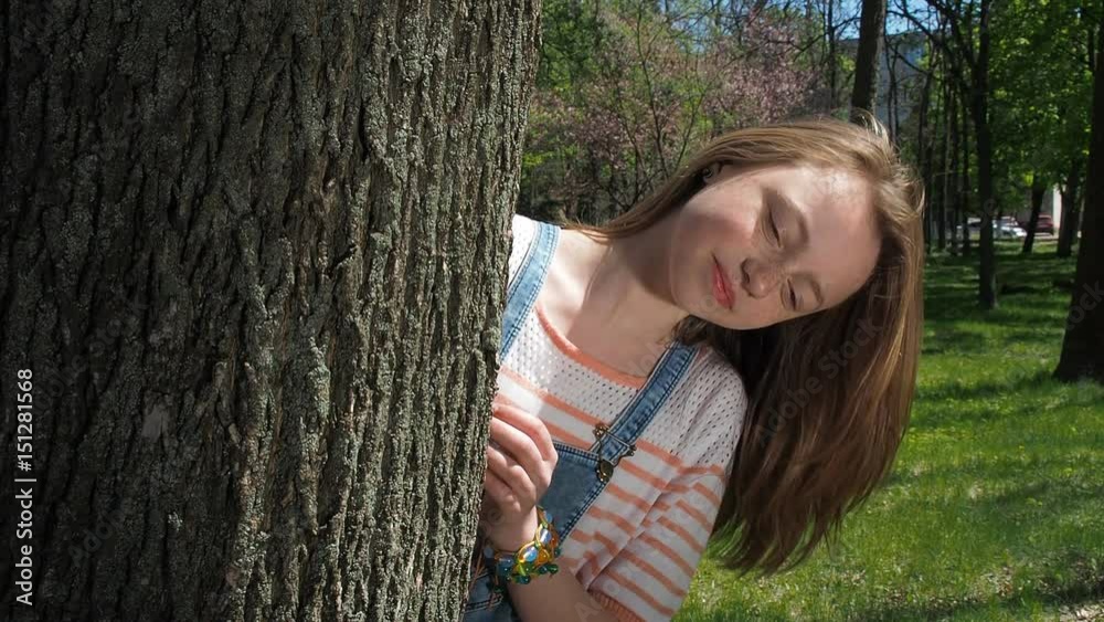 A girl with freckles looks out from behind a tree. Beautiful teen girl in the park. Emotion of the girl. A girl with freckles looks out from behind a tree. Beautiful teen girl in the park. Emotion of the girl.