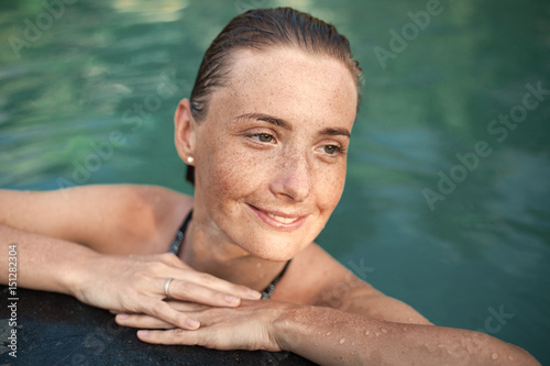 Horizontal close-up portrait of beautiful girl with sunspots and wet hair in water, arms on pool edge
