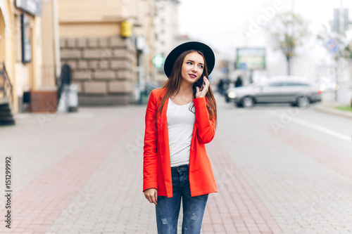 Pretty young Woman in black hat and red jacket using mobile phone on city street urban background. girl talking on the phone