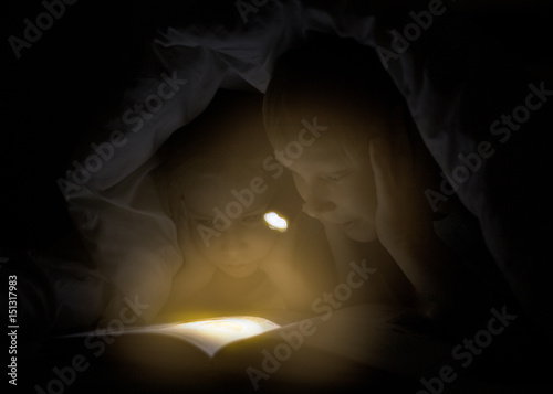 Children bedtime. Sister and brother are reading a book under a blanket with lashlight. Pretty young boy and lovely girl having fun in children room.