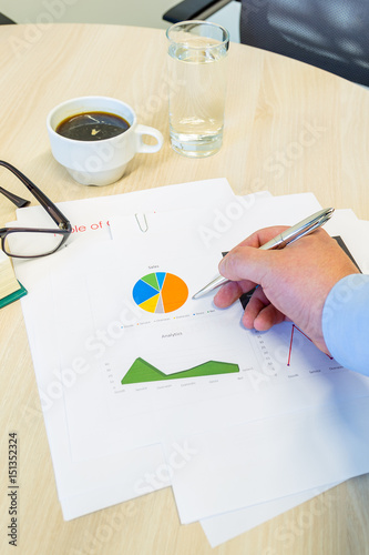 Businessman analyzing a pie chart and pointing his silver pen. His coffee, glasses and water are on the wood table.