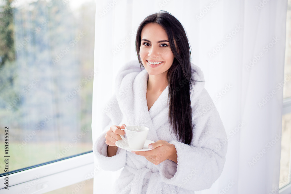 Nice beginning of day! Portrait of gorgeous brunette girl in bathrobe standing near window. She is smiling and holding a cup of coffee Nice beginning of day! Portrait of gorgeous brunette girl in bathrobe standing near window. She is smiling and holding a cup of coffee