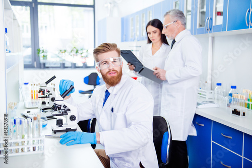 Portrait of young handsome bearded stylish intern, who is wearing gloves and labcoat, safety glasses. Behind him are people who are working for same experiment