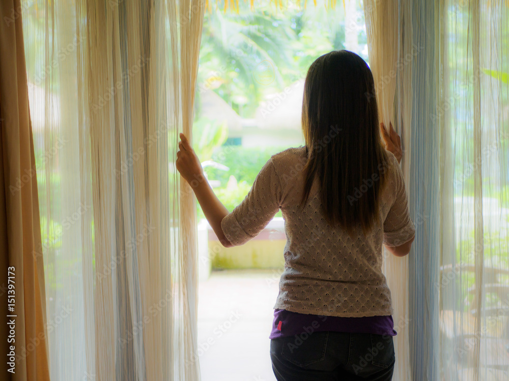 Rear view of a young woman holding the curtains open to look out of a large light window at home, interior. Positive and aspirational lifestyle. Woman looking out a window, indoors.