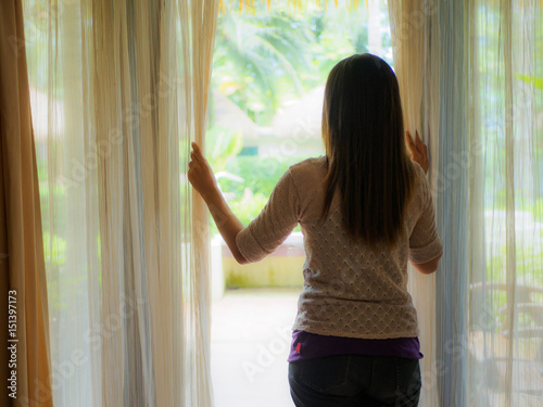 Canvastavla Rear view of a young woman holding the curtains open to look out of a large light window at home, interior