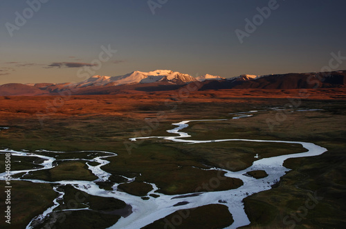 Snowcapped high ice mountain peaks with glaciers above orange steppe with whitewater river under dramatic dawn sky. Altai Plato Ukok Siberia Russia