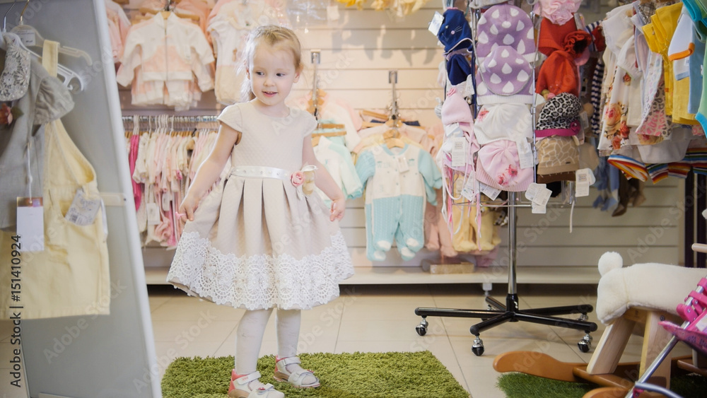 Blonde little girl chooses new dress near mirror in store of kids clothes