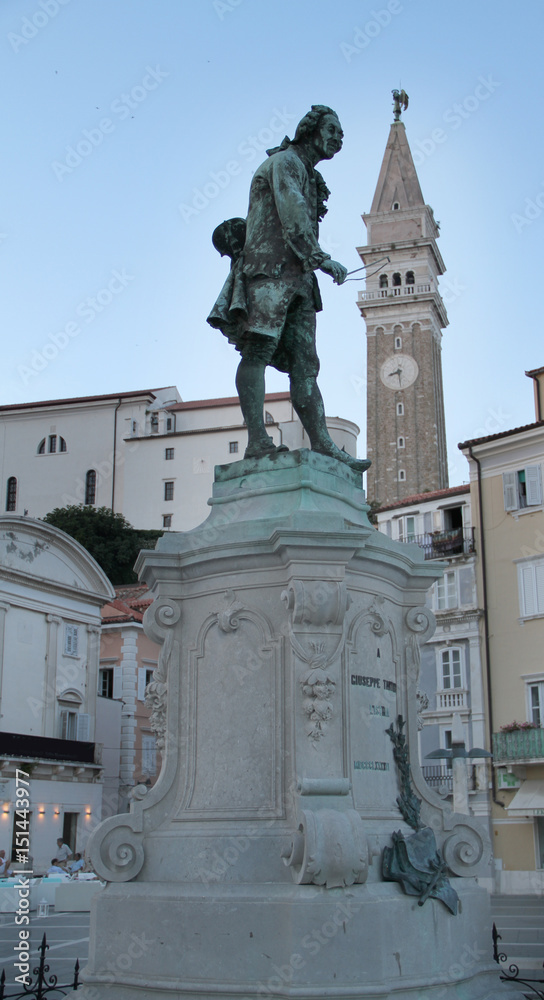 A main square in the town of Piran. A statue of a musician in the front and an old church in the background.