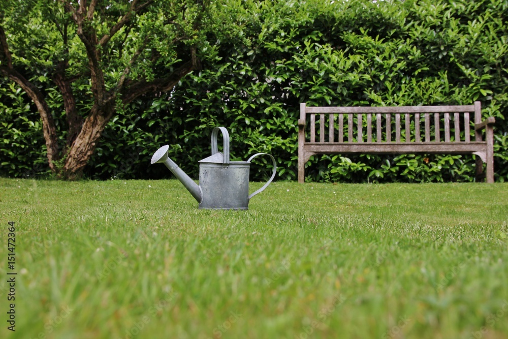 Steel watering can and wooden bench in an italian garden Steel watering can and wooden bench in an italian garden