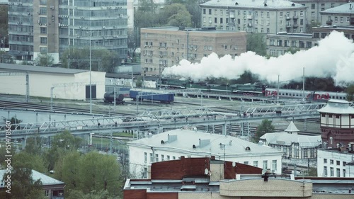 View from distance of big railway station and old vintage locomotive blowing a lot of smoke while passing through residential district, puffer on railroad during Victory's day in Moscow, Russia