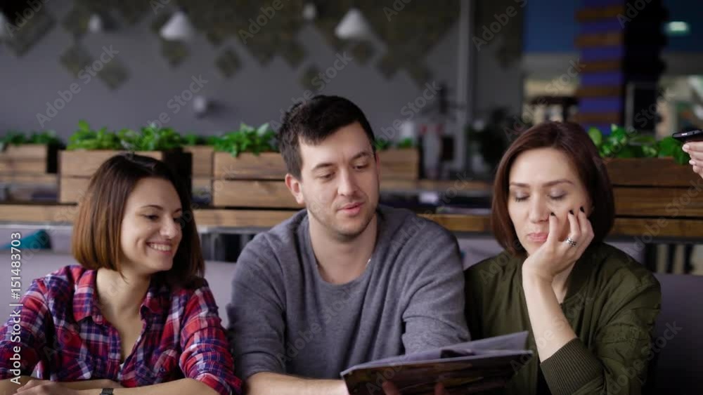 A waiter girl who wears an apron accepts an order from three friends with dark hair who sits in a restaurant and choose dishes from the menu A waiter girl who wears an apron accepts an order from three friends with dark hair who sits in a restaurant and choose dishes from the menu