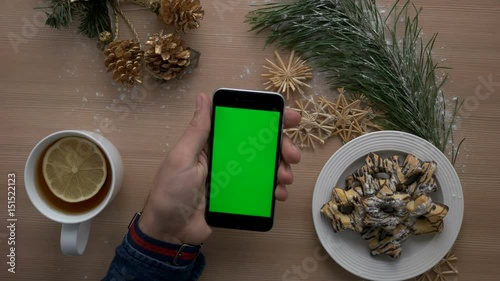 Man using smart phone with green screen on wooden table with christmas decoration. Cozy christmas details. Top view. Chroma key photo