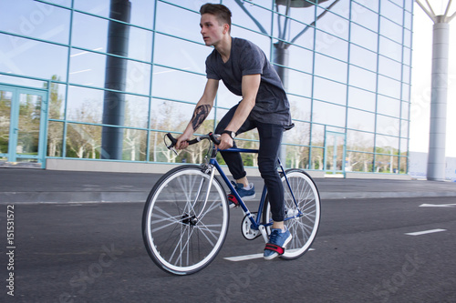  portrait of a young man riding on bicycle in city street. Man on blue bicycle with white wheels, big mirror windows background