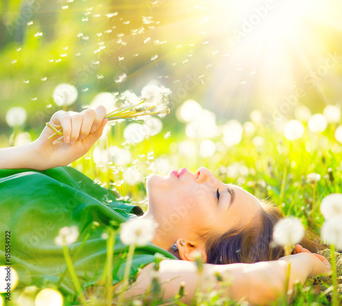 Beautiful young woman lying on the field in green grass and blowing dandelion