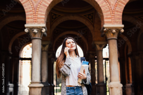 Gorgeous happy student holding notebooks speak phone on campus at college