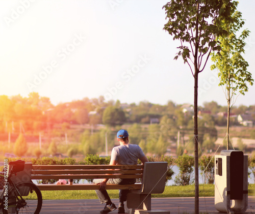 Lonely young man sitting on a bench in the park near the lake on sunset, coffee break