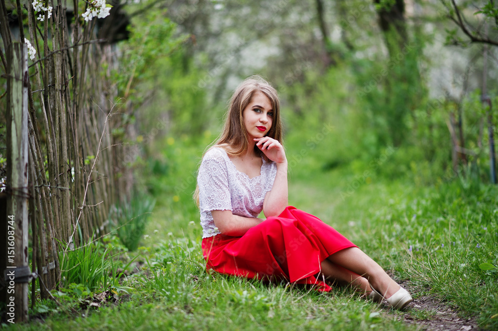 Portrait of sitting beautiful girl with red lips at spring blossom garden on green grass, wear on red dress and white blouse. Portrait of sitting beautiful girl with red lips at spring blossom garden on green grass, wear on red dress and white blouse.