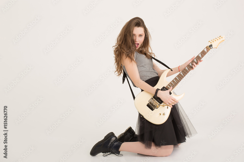 Hot girl emotionally playing the electric guitar standing on her knees isolated. Brunette model playing hard rock music and singing at camera isolated. Sexy musician girl with dark lipstick