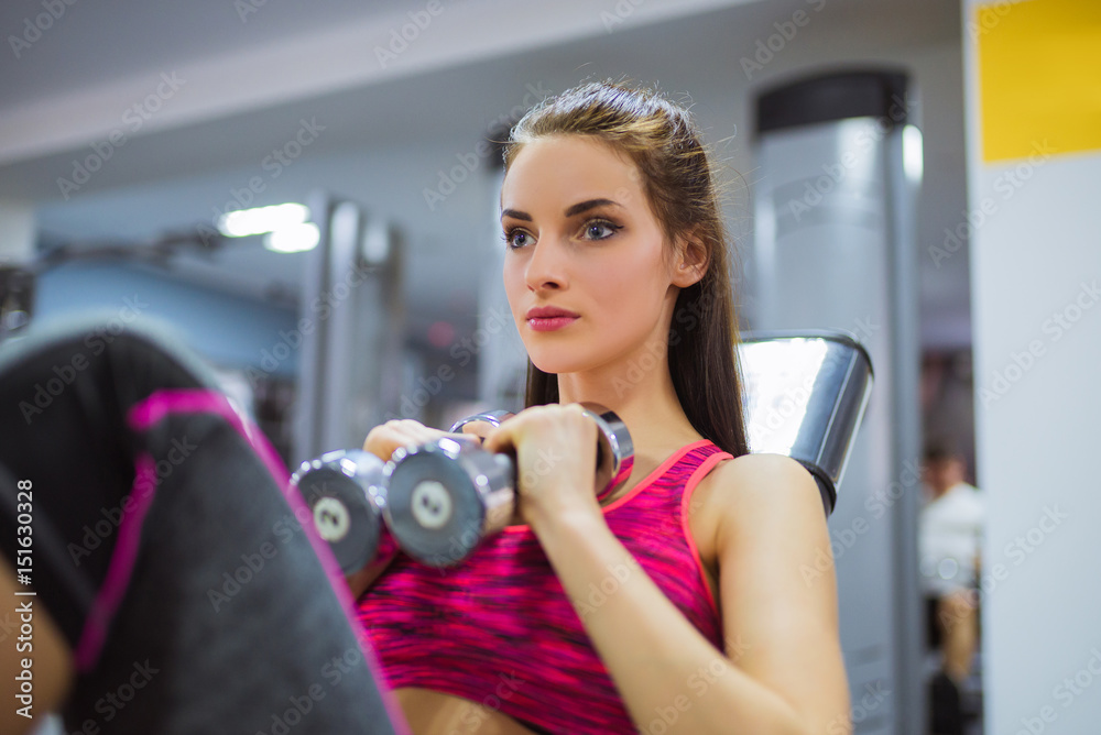 Close-up focused sports woman sits on simulator and keeps dumbbells in hands, at the gym