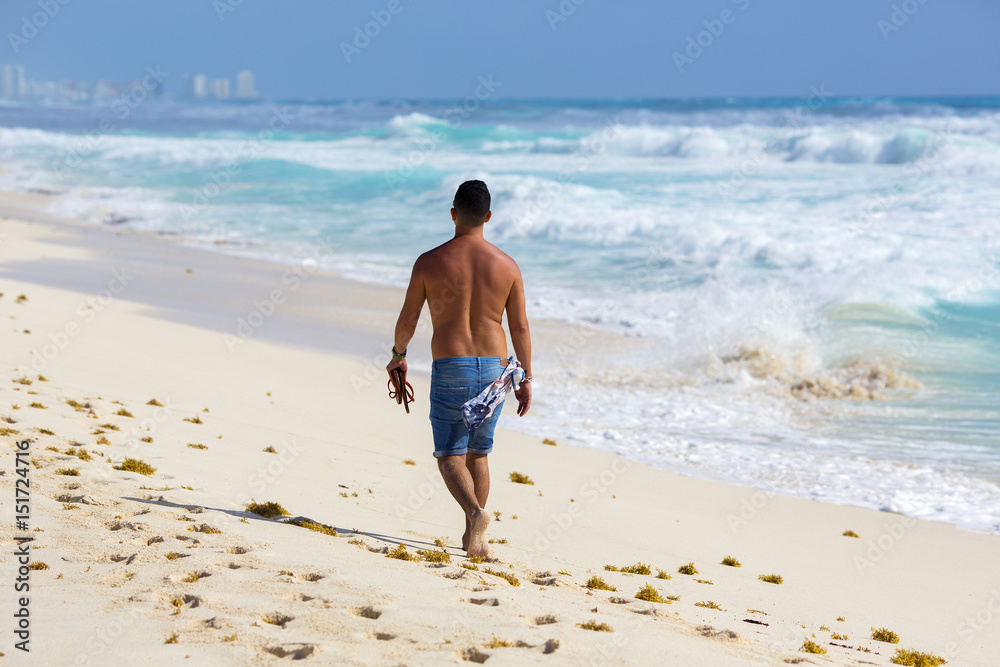 Young man is walking the beach shirtless. Barefoot walk on a sunny day. The shore of the Caribbean sea.