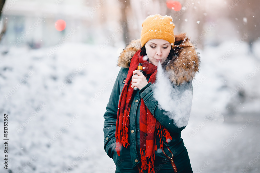 Fototapeta premium girl is holding an electronic cigarette in her hand and exhaling steam from her mouth. She is wearing a hat. Winter. Concept of safe smoking, giving up tobacco. Toning, highlighting