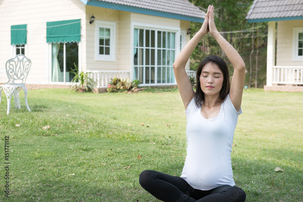 asian pregnant woman practicing yoga on green grass in front of her house. meditating on maternity. concept of healthy lifestyle and relaxation.