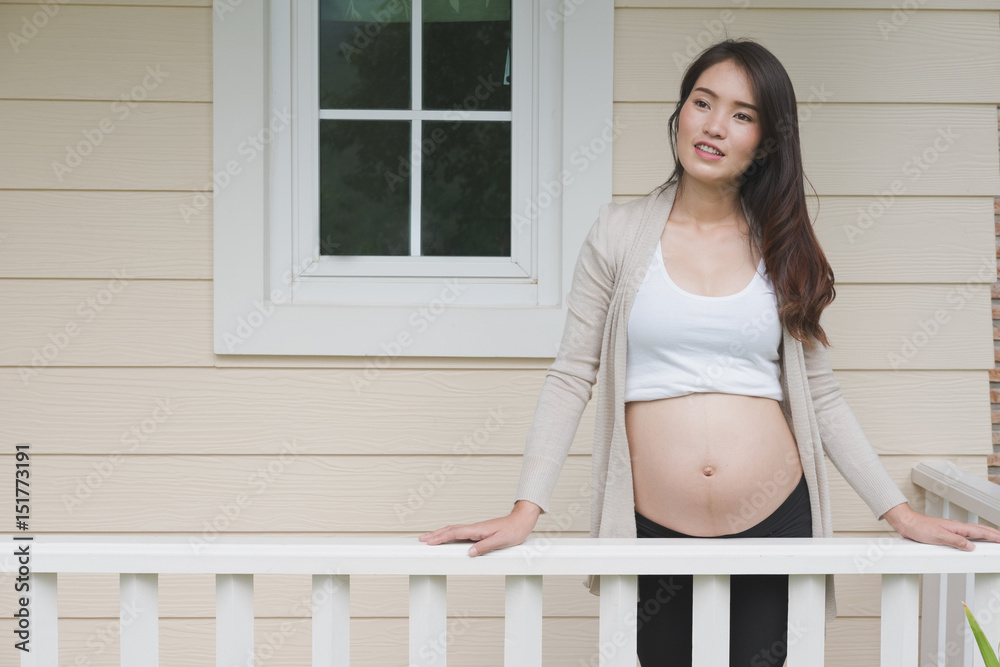 Young asian woman standing on balcony smiling with pleasure. Beautiful female expecting child having rest in front of her house Young asian woman standing on balcony smiling with pleasure. Beautiful female expecting child having rest in front of her house