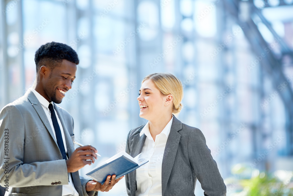 Portrait of two young business colleagues, one of them African, discussing work smiling cheerfully standing in glass hall of modern office building Portrait of two young business colleagues, one of them African, discussing work smiling cheerfully standing in glass hall of modern office building