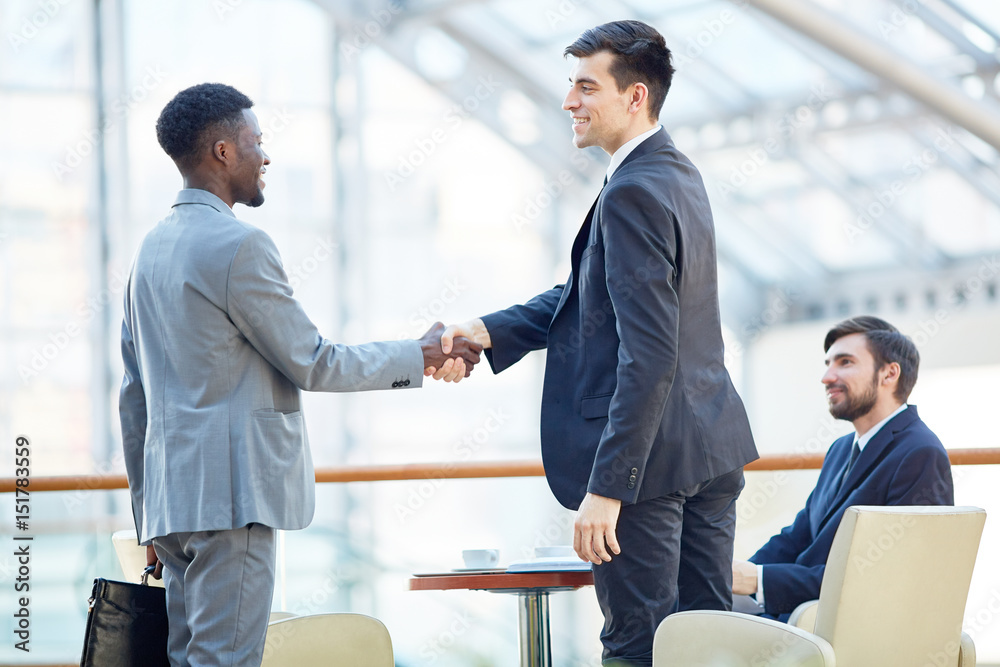 Portrait of smiling successful businessman greeting African-American man in meeting shaking hands at table in office building Portrait of smiling successful businessman greeting African-American man in meeting shaking hands at table in office building