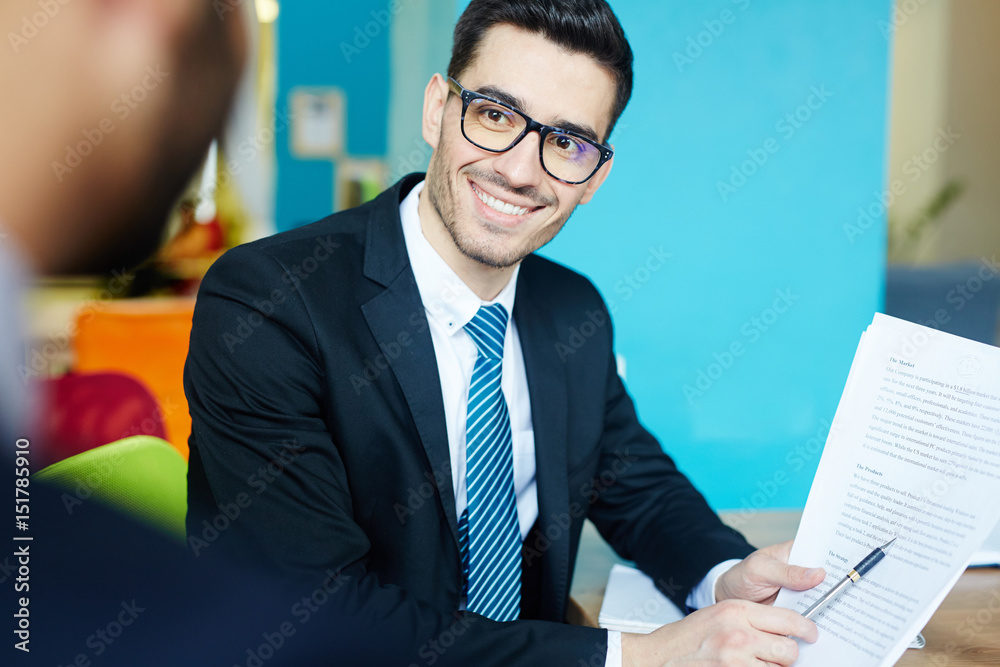 Happy young businessman pointing at document in his hand while discussing it with colleague