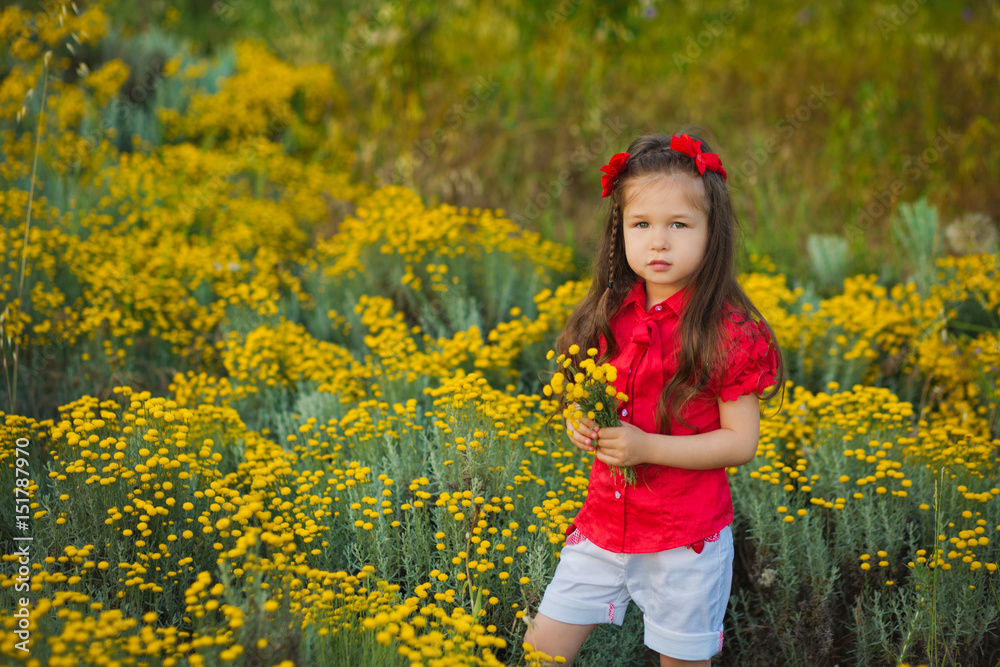 Young cute girl in red shirt and white shorts standing close to yellow flowers.