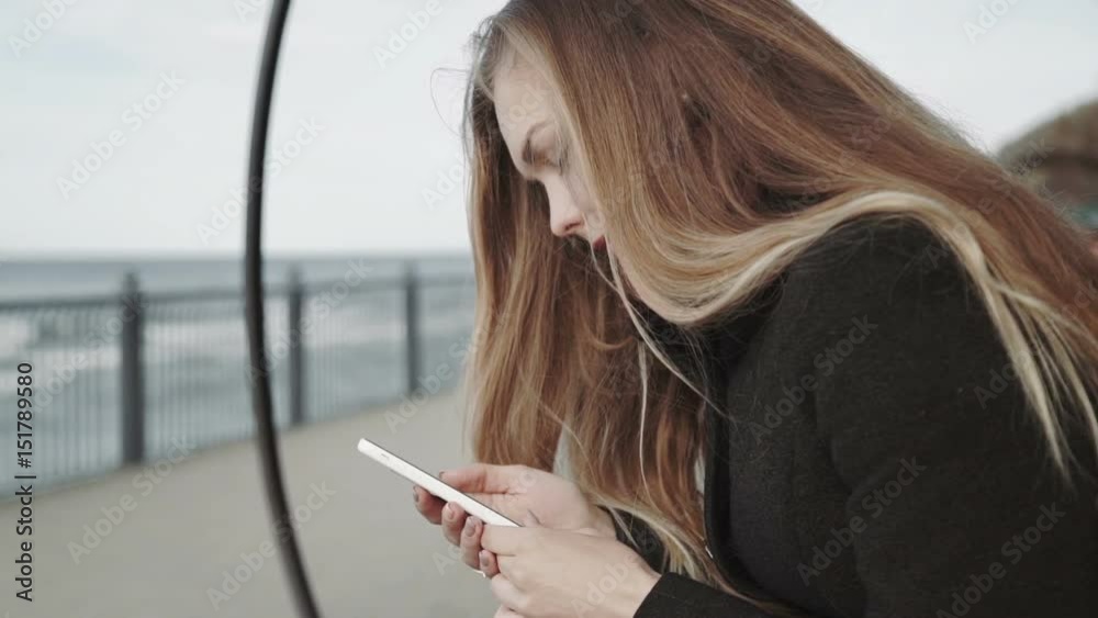 Woman with white smartphone on the bench on the boardwalk overlooking the sea. Woman with white smartphone on the bench on the boardwalk overlooking the sea.
