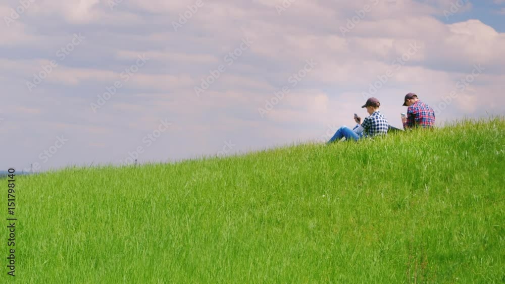 Two teenagers use mobile phones in a picturesque place on a green hill. A serene landscape - technology and be in touch anywhere. Copyspace composition Two teenagers use mobile phones in a picturesque place on a green hill. A serene landscape - technology and be in touch anywhere. Copyspace composition