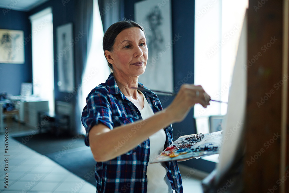 Fototapeta premium Portrait of elegant mature woman painting oil picture on canvas in art-studio, looking inspired and enjoying process