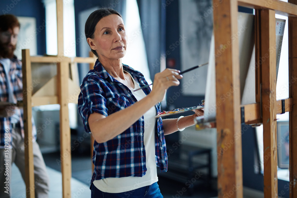 Fototapeta premium Portrait of inspired mature woman painting picture on easel standing in row of students in art class