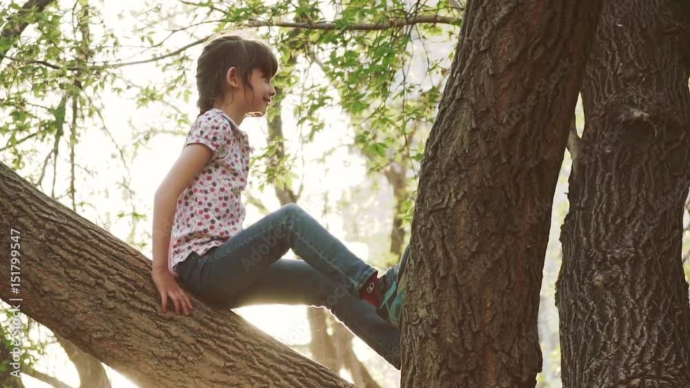 child is playing in the park in the summer. Little girl is sitting on a tree branch. Adventure of a children. Happy carefree childhood. slow motion