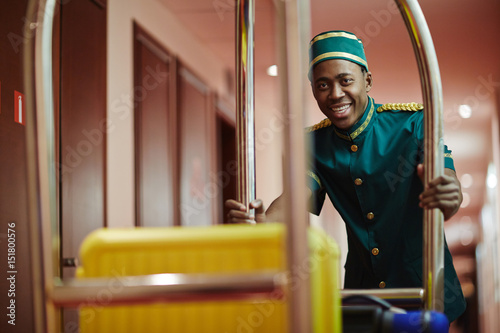 Portrait of smiling African bellhop helping guests, pushing luggage cart delivering bags to hotel rooms