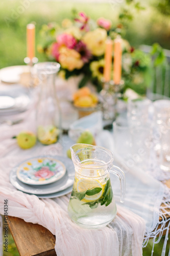picnic, food, summer, holiday concept - full glass jug of fresh lemonade on festive table, slices of lemon, lime, mint leaves in flagon, bouquet, candles, glasses and plates, fruits, selective focus