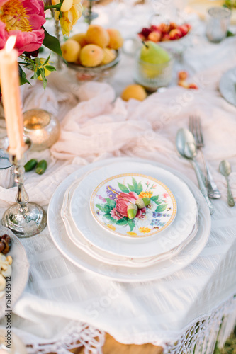 picnic, food, holiday concept - holiday decorated table with white tablecloth, stack of white plates with floral pattern, two acorns on top, burning candle in candlestick, flowers, plates with fruits