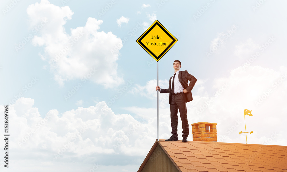 Young businessman on house brick roof holding yellow signboard. Mixed media Young businessman on house brick roof holding yellow signboard. Mixed media