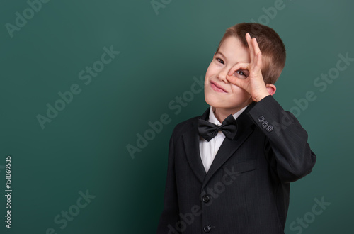 school boy show ok sign, portrait near green blank chalkboard background, dressed in classic black suit, one pupil, education concept