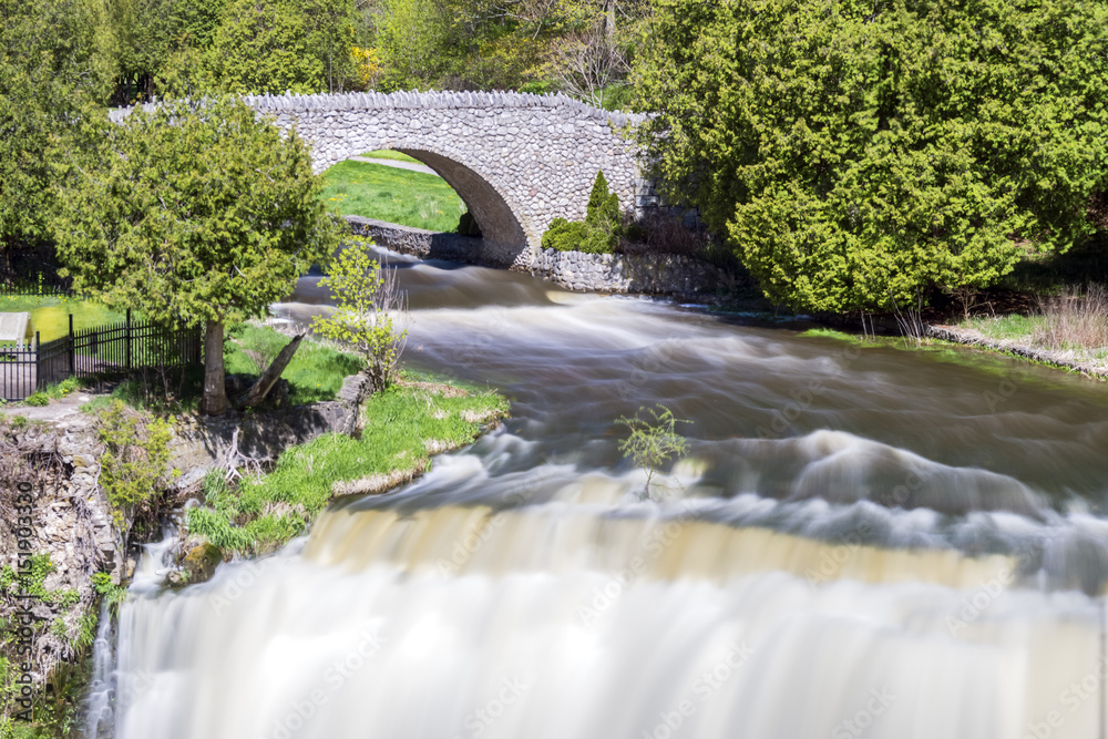 Fototapeta premium Stone bridge over river bend and waterfall, smooth flowing current